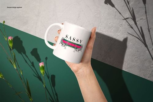 Hand holding a white 11oz mug with a floral and text design, shown against a green and gray background with pink flowers.