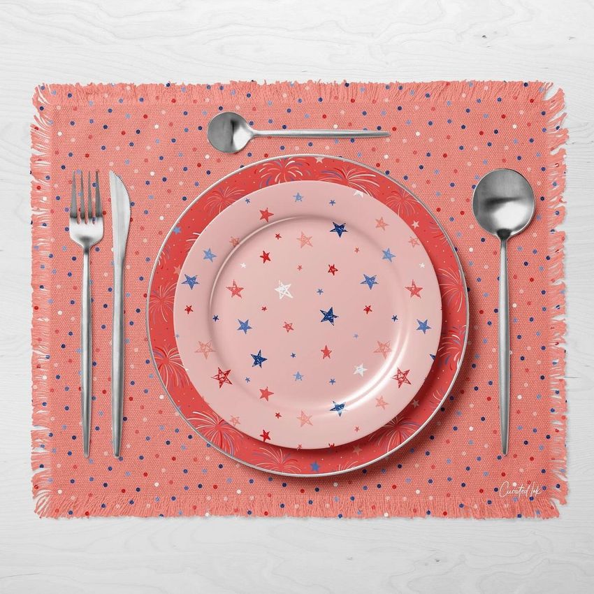 Fringed placemat mockup with a red and pink star-patterned plate set, silverware, and spoon on a light wood surface.