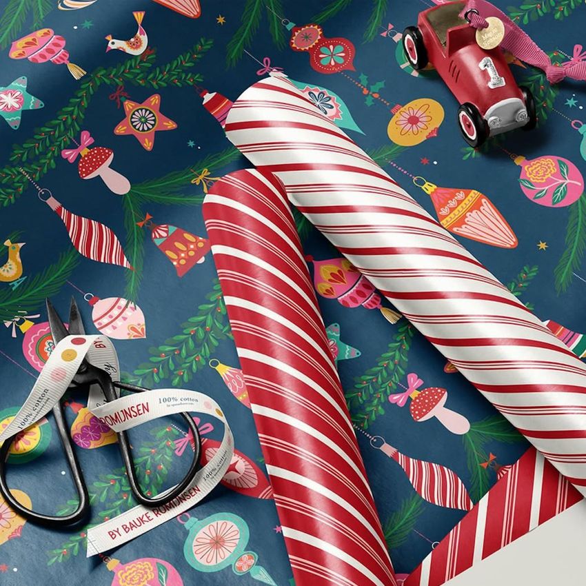 Gift wrapping paper mockup showing festive Christmas patterns with ornaments and birds, plus candy cane striped rolls and scissors.