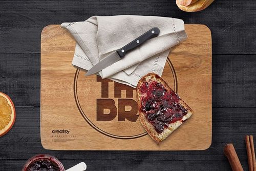 Rectangular engraved wooden board mockup with bread and jam, knife, and linen napkin on a dark table surface.