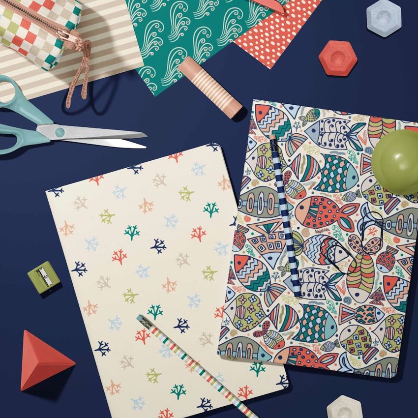 Stationery mockup featuring two A4 notebooks with colorful sea-inspired patterns, surrounded by patterned papers and office supplies on a dark blue surface.