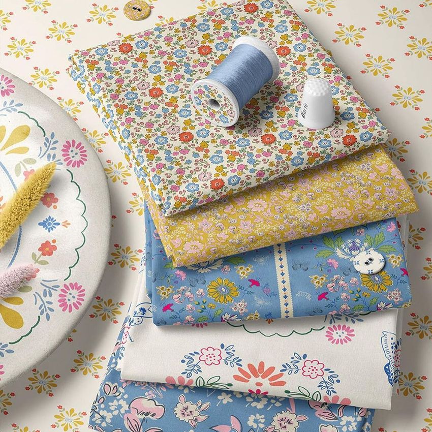 Fabric stack mockup featuring five folded fabrics with floral patterns in pastel colors, shown with a spool of blue thread and a thimble on a patterned surface.