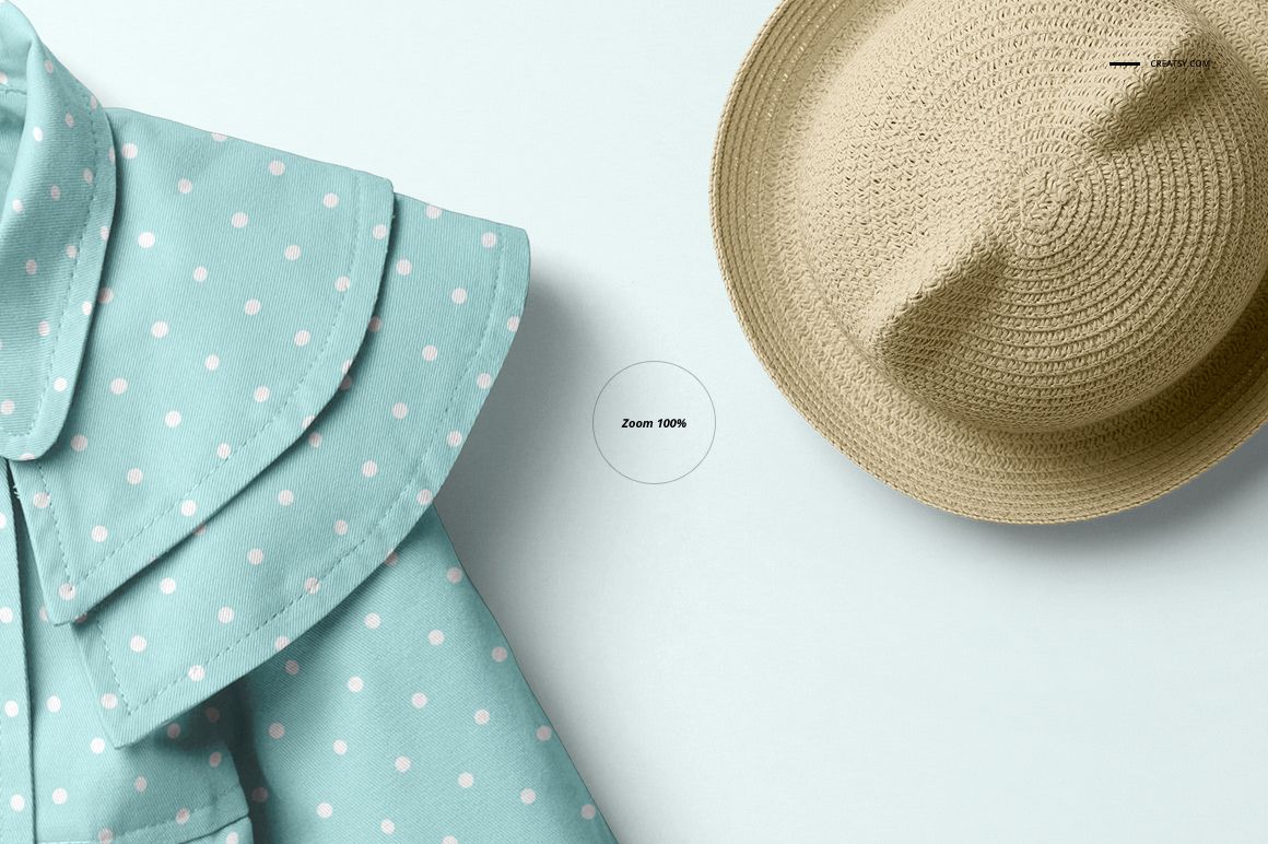 Front view of a baby coat mockup with light blue fabric and white polka dots, next to a beige straw hat on a pale background.