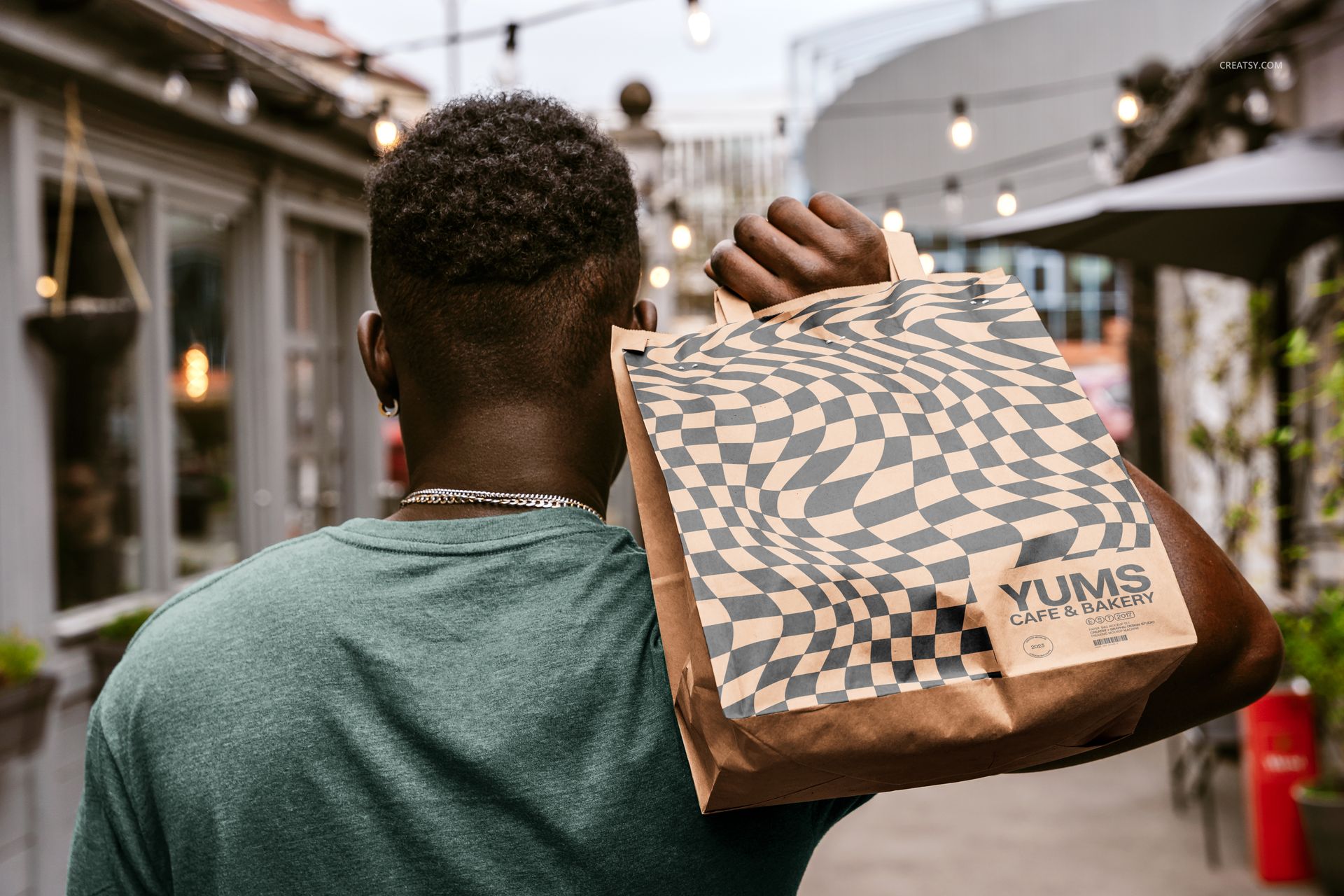 Brown paper bag mockup with bold geometric design, carried by a person in an outdoor alleyway with string lights.