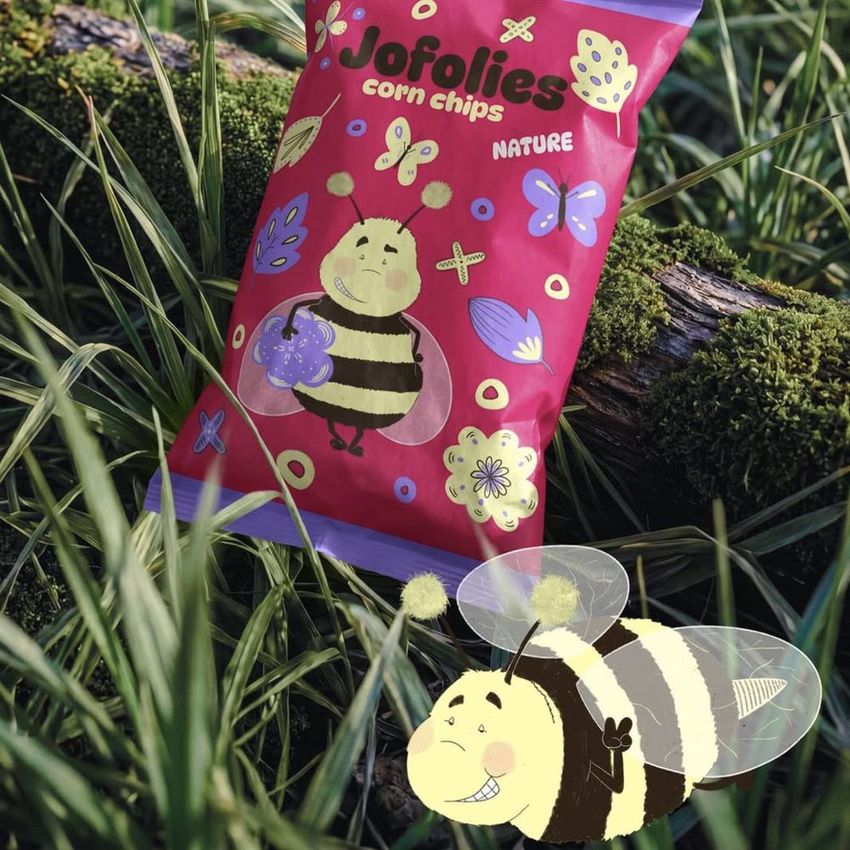 Paper chips bag mockup featuring a whimsical bee illustration and colorful nature motifs, placed on a mossy log in tall grass.