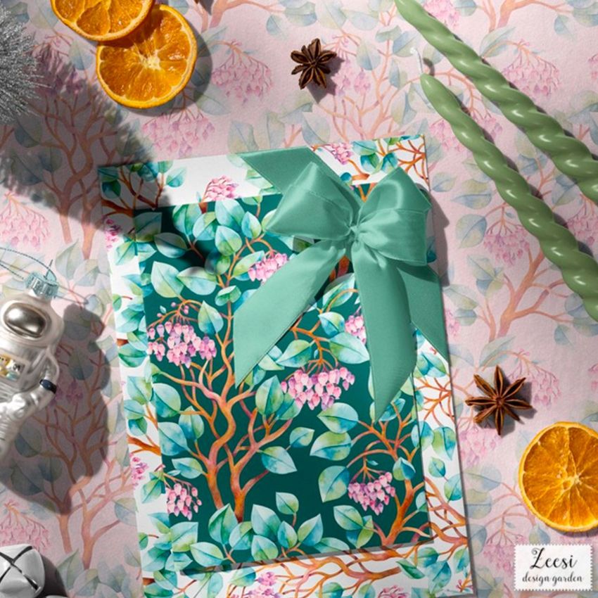 Christmas greeting card mockup featuring a watercolor floral design with green leaves and pink flowers, topped with a teal ribbon bow on a patterned surface.