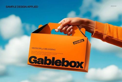PSD mockup showing a bright orange gable box packaging held up outdoors, with clouds and blue sky in the background.