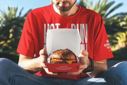 PSD mockup showing a man holding a burger box with a sandwich, outdoor setting featuring lush palm trees.