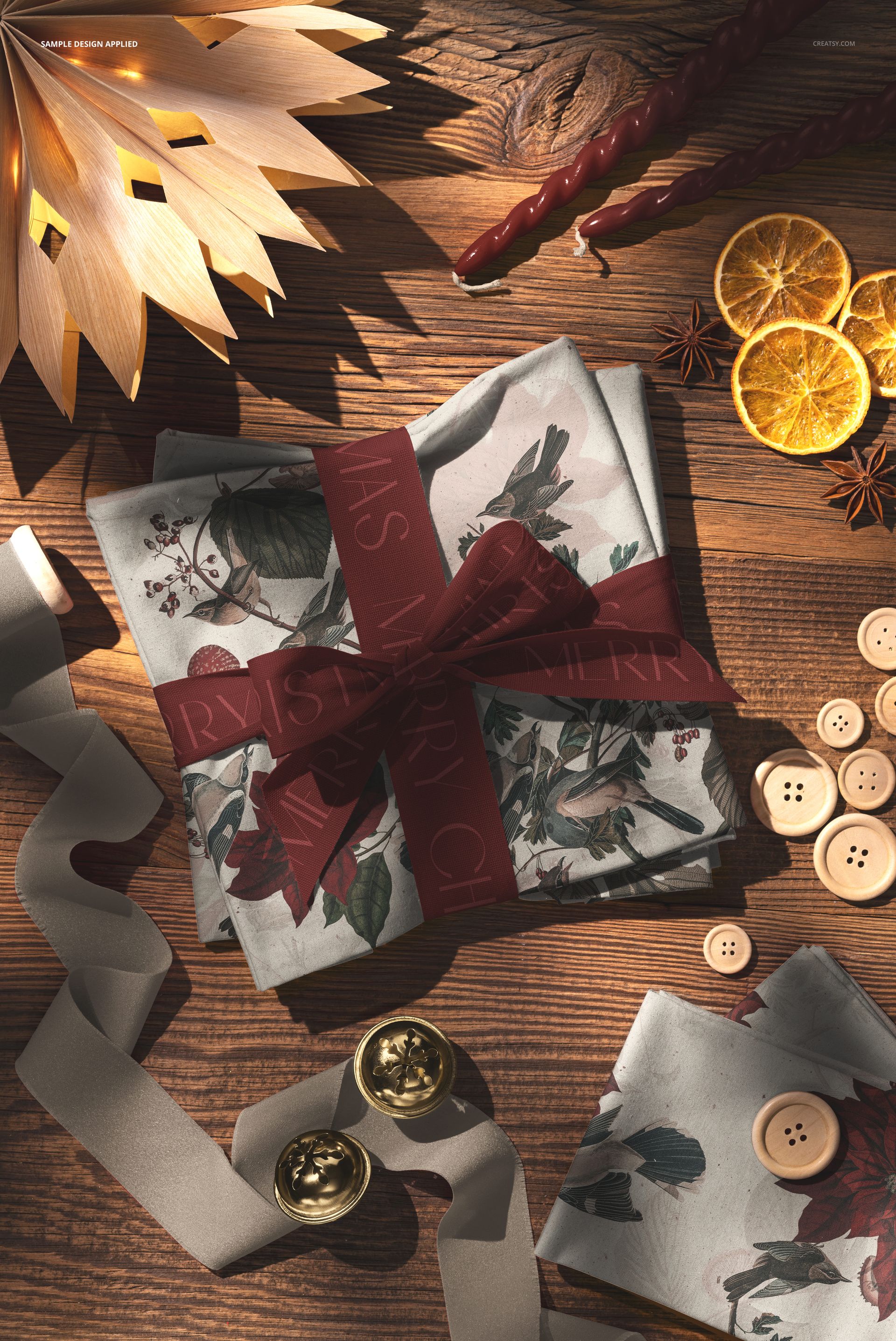 Christmas-themed folded cotton fabric mockup on a wooden surface, surrounded by buttons, ribbon, and dried orange slices.