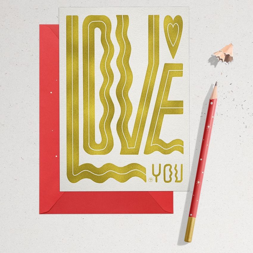 Stationery card mockup featuring a white card with bold gold text and a heart, placed on a red envelope with a pencil and pencil shavings nearby.