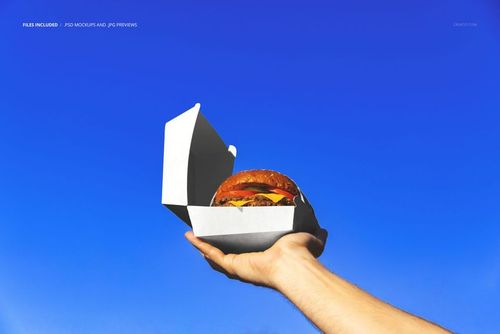 Hand holding an open white burger box with a hamburger inside against a clear blue sky, front view mockup.