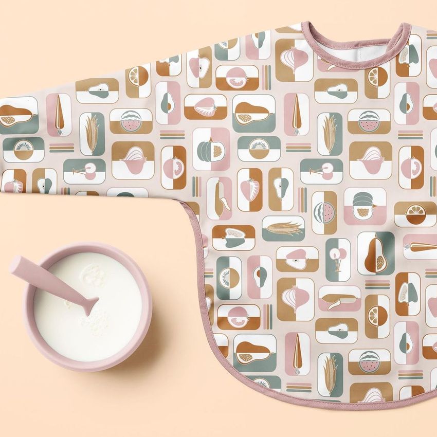 Baby sleeved bib mockup featuring a colorful geometric food pattern, shown flat on a beige surface beside a bowl of milk.
