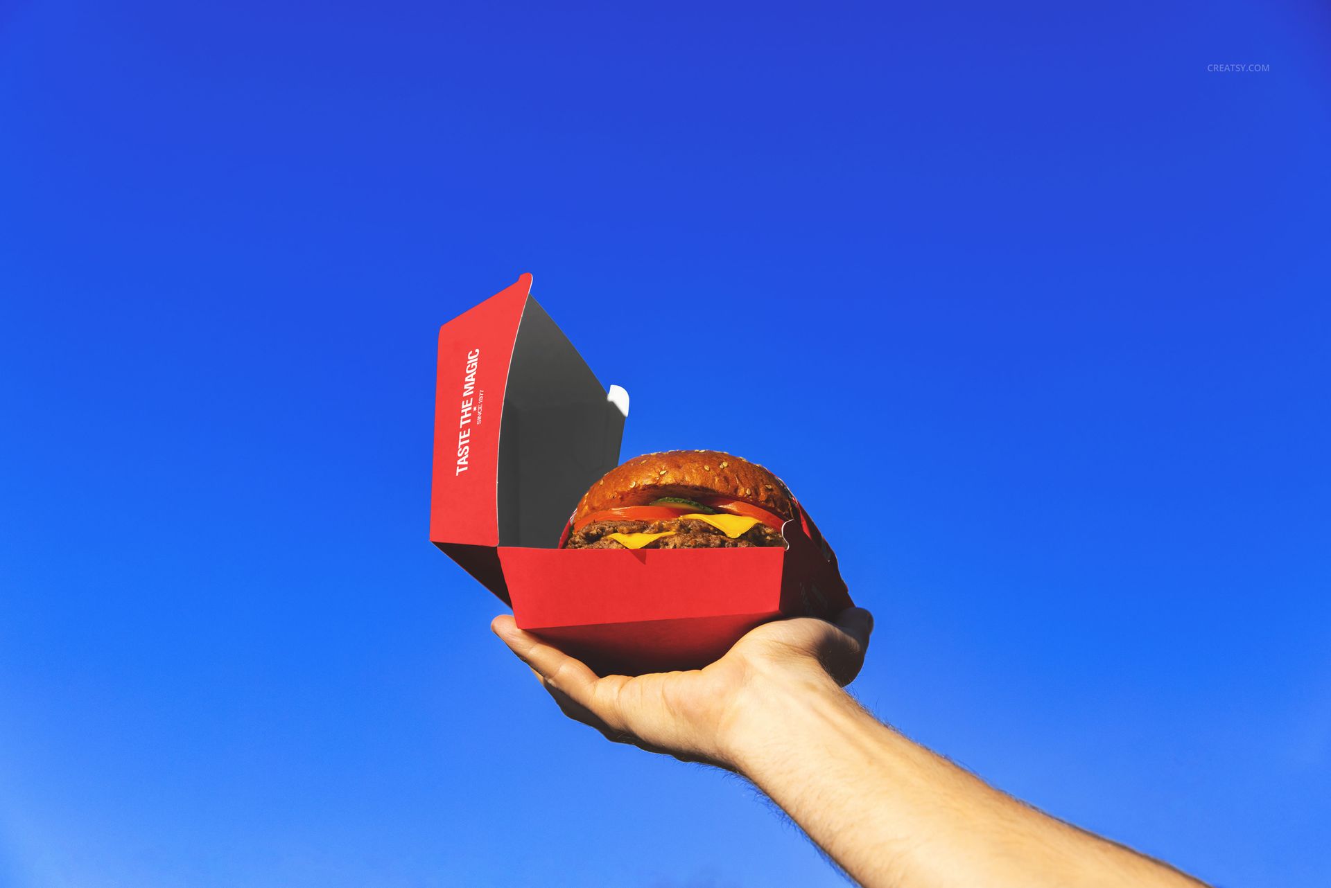 Open red burger packaging mockup held up by a hand, featuring a cheeseburger and bright blue sky backdrop.