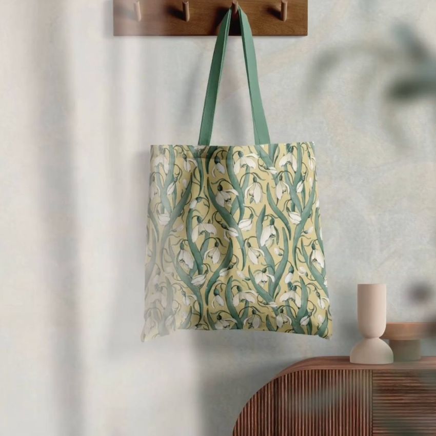 Tote bag mockup hanging on a wooden hook, featuring a floral snowdrop pattern with green handles and a soft neutral background.