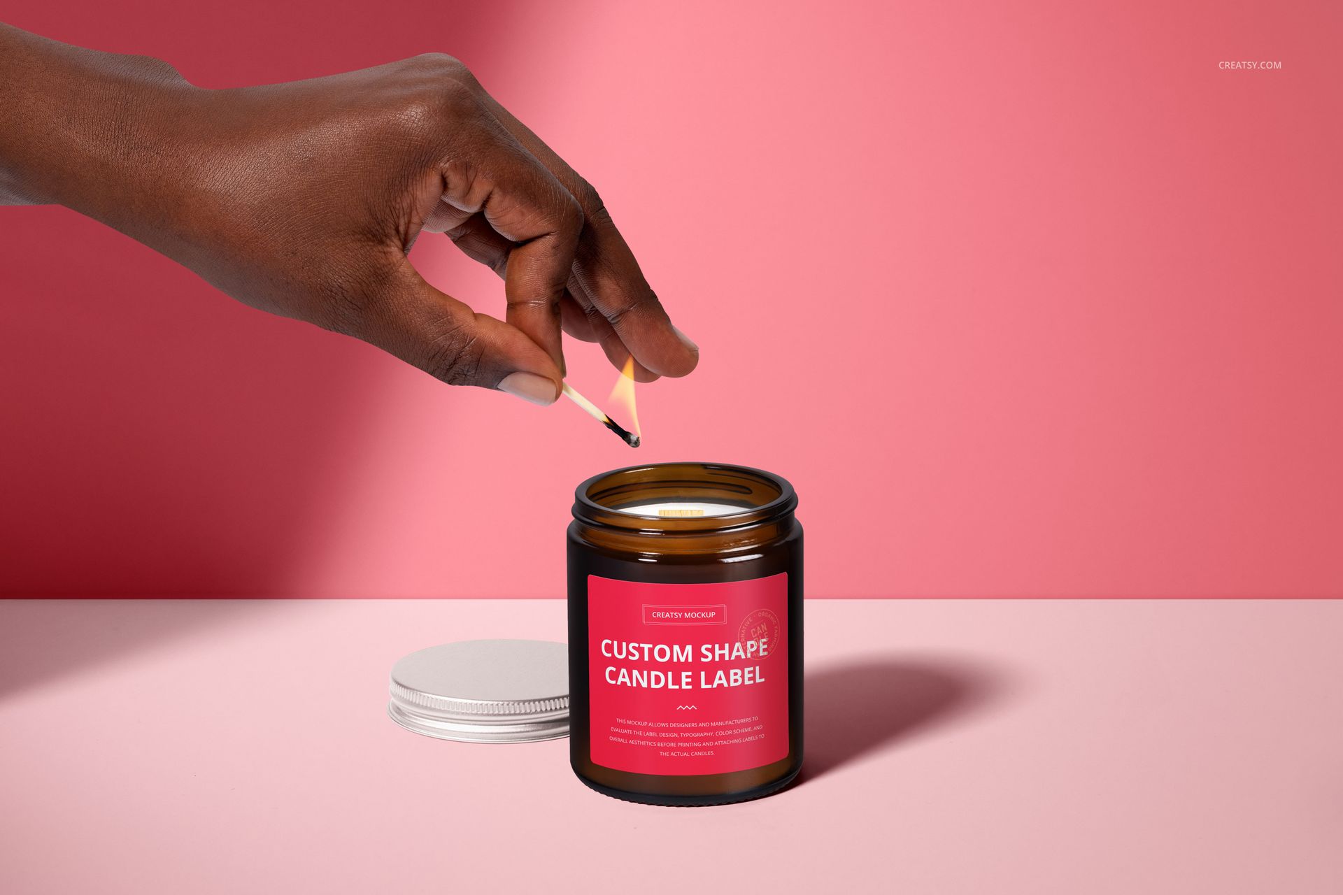 PSD mockup of amber candle jar with wooden wick, hand holding match above jar, pink backdrop and silver lid nearby.