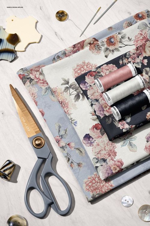 PSD mockup featuring layered floral jersey fabrics, sewing scissors, threads, needles, and buttons on a table.