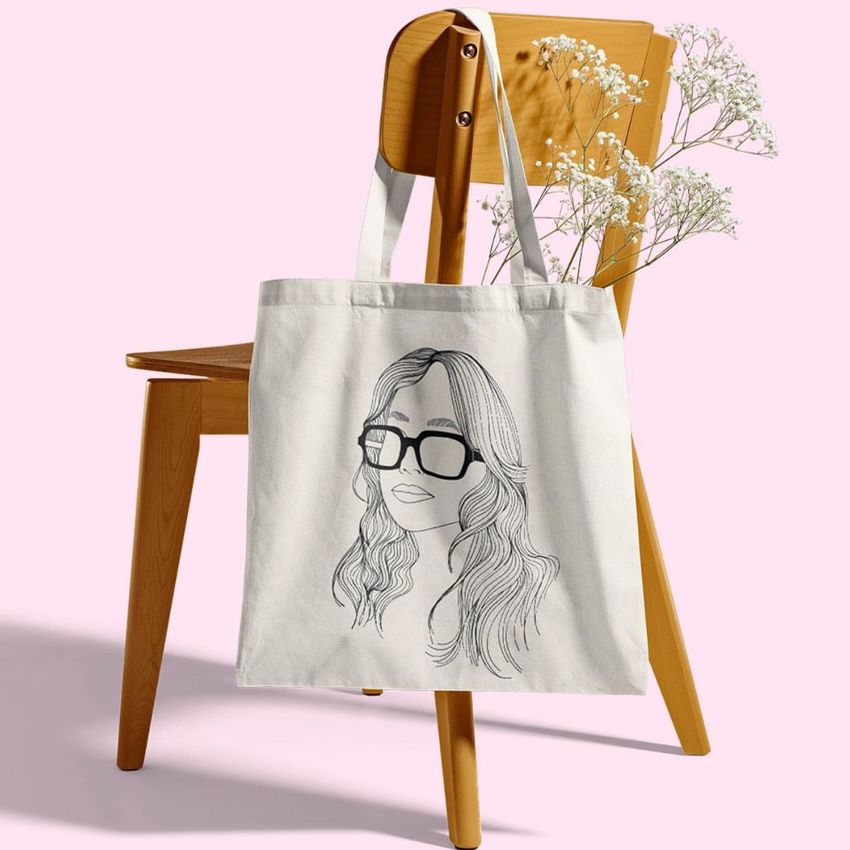 Tote bag mockup displayed on a wooden chair with a floral illustration, set against a pale pink background with white flowers.