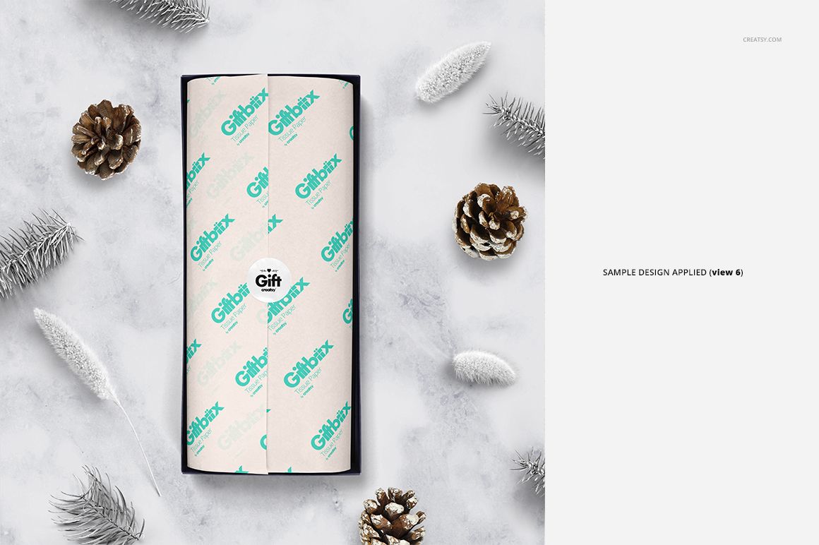 Gift box mockup featuring white tissue paper with teal lettering, surrounded by pinecones and frosted branches, top view.