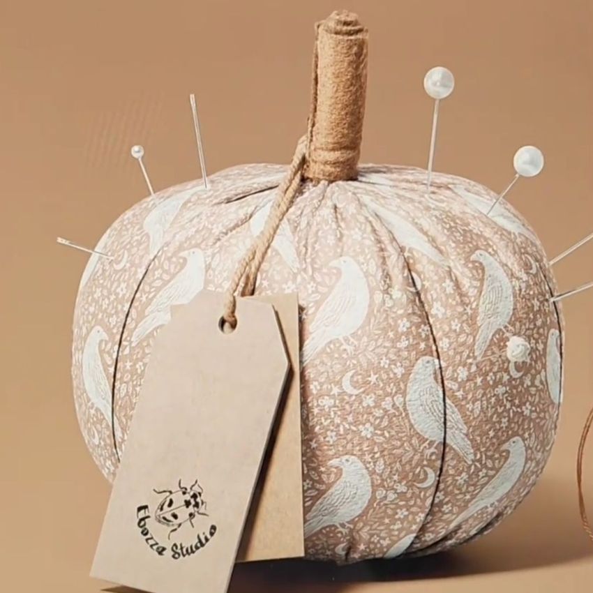 Fabric pumpkin mockup featuring a beige pumpkin with white bird and floral patterns, pins inserted on top, and a kraft tag attached.
