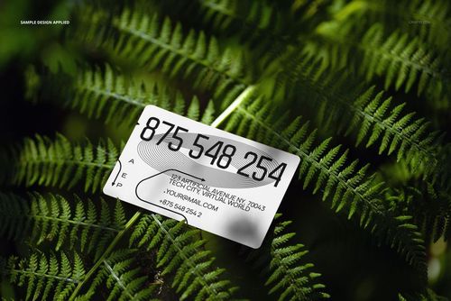 PSD mockup of a business card with simple black typography displayed on vibrant green fern leaves outdoors.