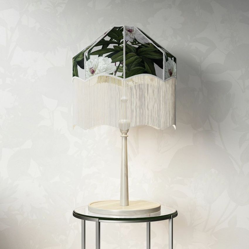 Table lamp mockup with a tasseled shade featuring a dark floral pattern, placed on a round marble-top table against a light floral wallpaper.