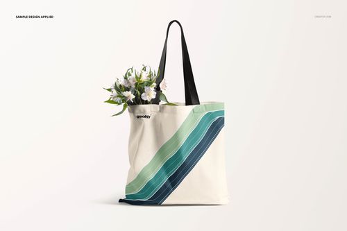 Tote bag mockup with a minimalist stripe pattern in green and blue, black straps, and white flowers inside.