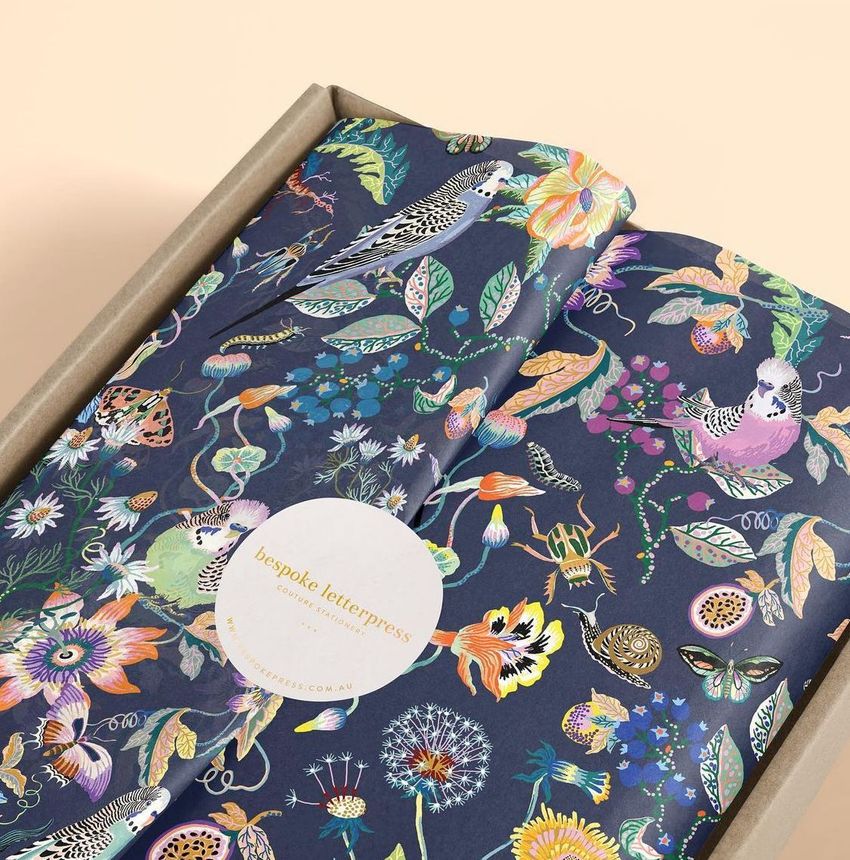 Mailer box mockup with dark blue wrapping tissue paper featuring a colorful budgie and floral pattern, photographed from above.