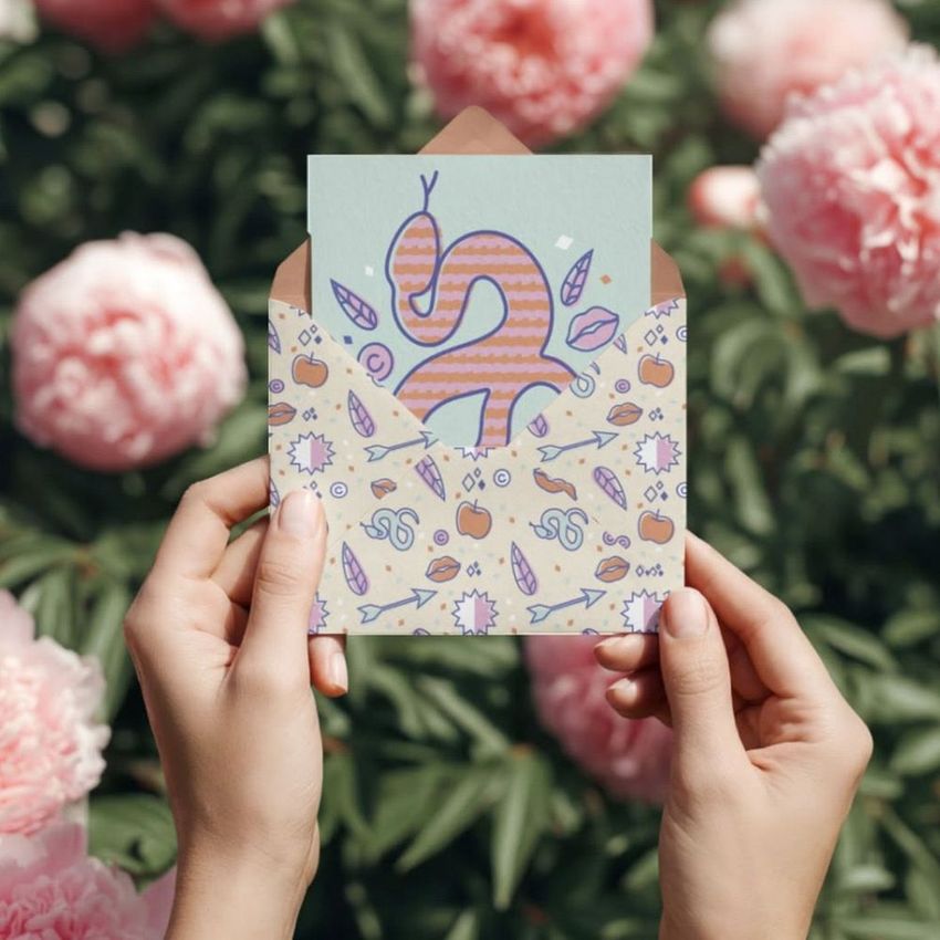 Hands holding an envelope and greeting card mockup with playful illustrated patterns, surrounded by blooming pink peonies outdoors.