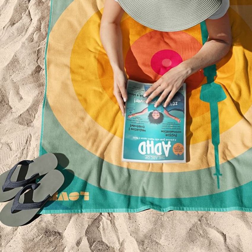 Beach towel mockup featuring a colorful geometric pattern, with a person reading a magazine and wearing a wide-brimmed hat on sand.