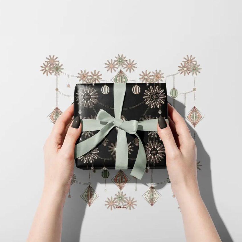 Hands holding a gift box mockup wrapped in black paper with metallic geometric patterns and a light green ribbon, on a white background.