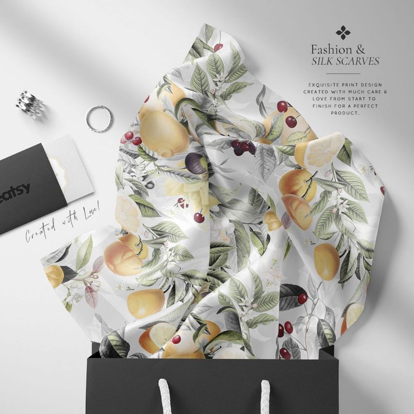 Silk scarf mockup with a botanical fruit pattern displayed draped over the edge of a black gift bag, set on a light background.