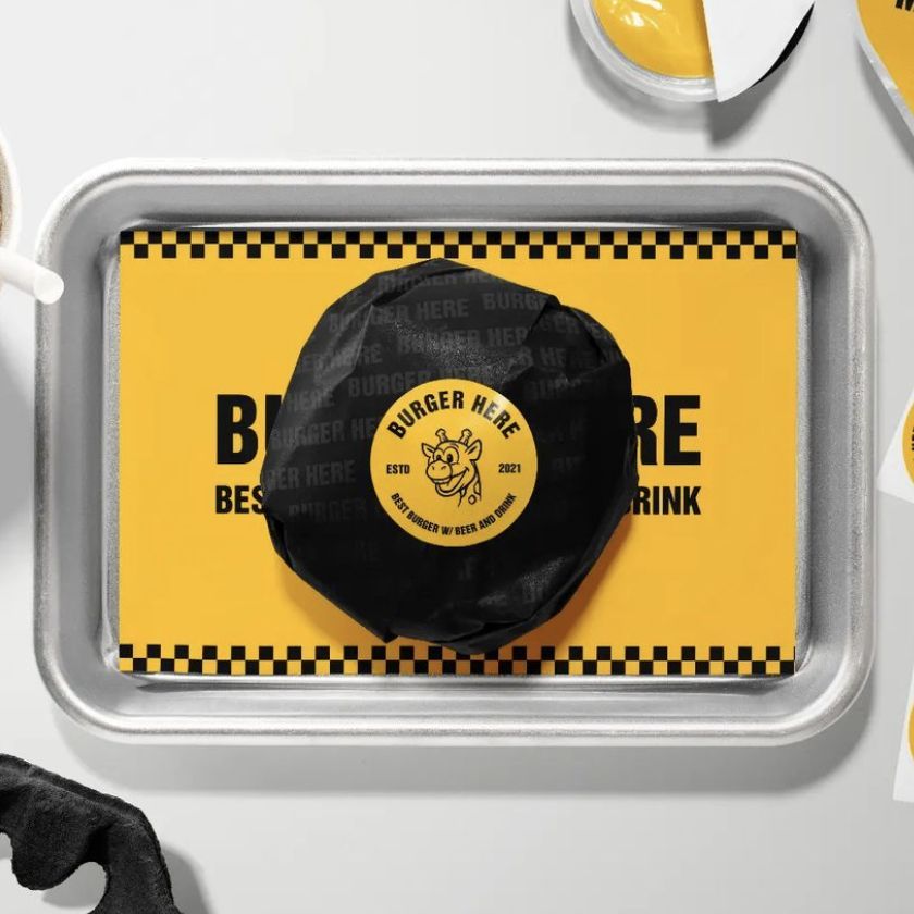Burger mockup featuring a black wrapped burger with text and a cartoon graphic, placed on a yellow and black checkered paper on a metal tray.