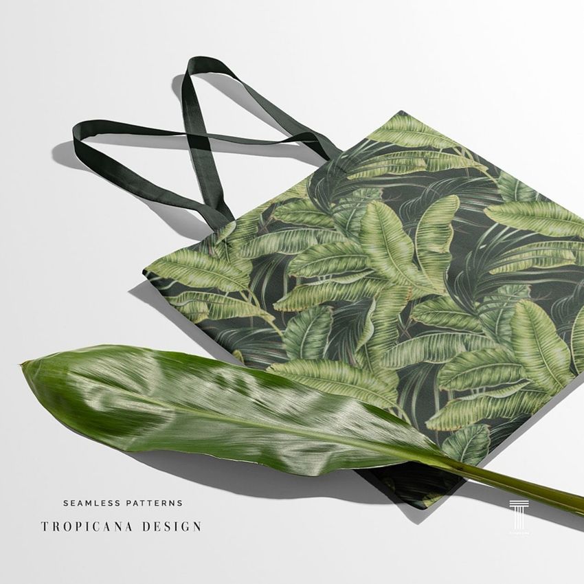 Tote bag mockup featuring a green tropical leaf pattern on a flat surface with a real leaf and soft shadows on a light background.