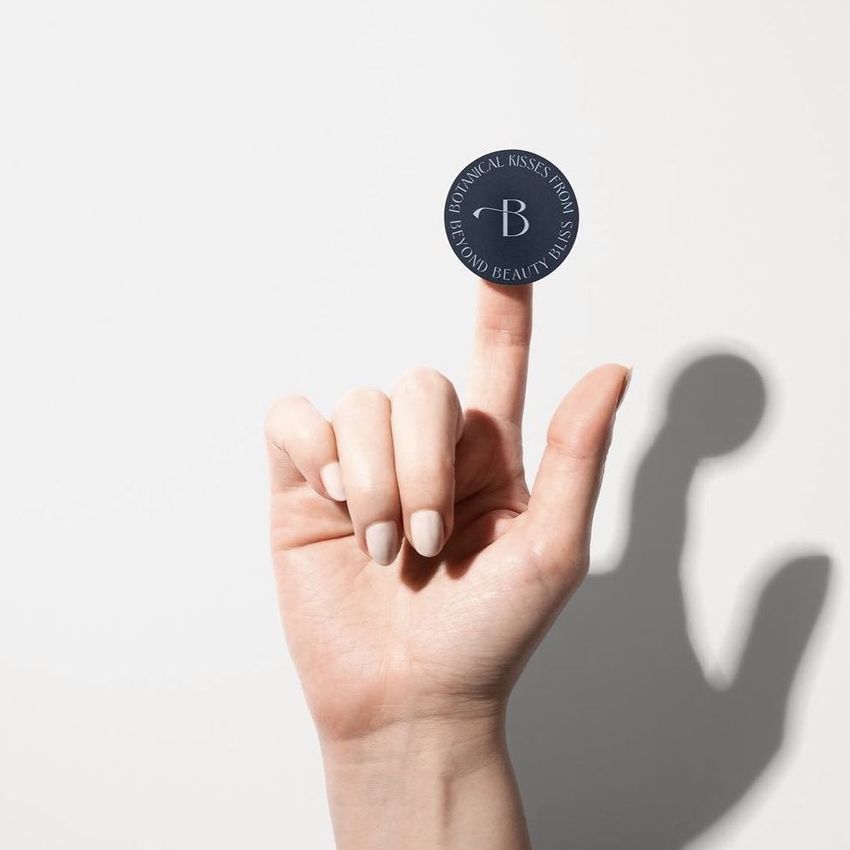 Hand with neutral nail polish holding a round dark sticker mockup on the index finger against a plain white background.