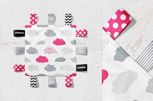 Square taggie blanket mockup with pink, gray, and white cloud pattern, surrounded by colorful fabric tags on a white wood surface.