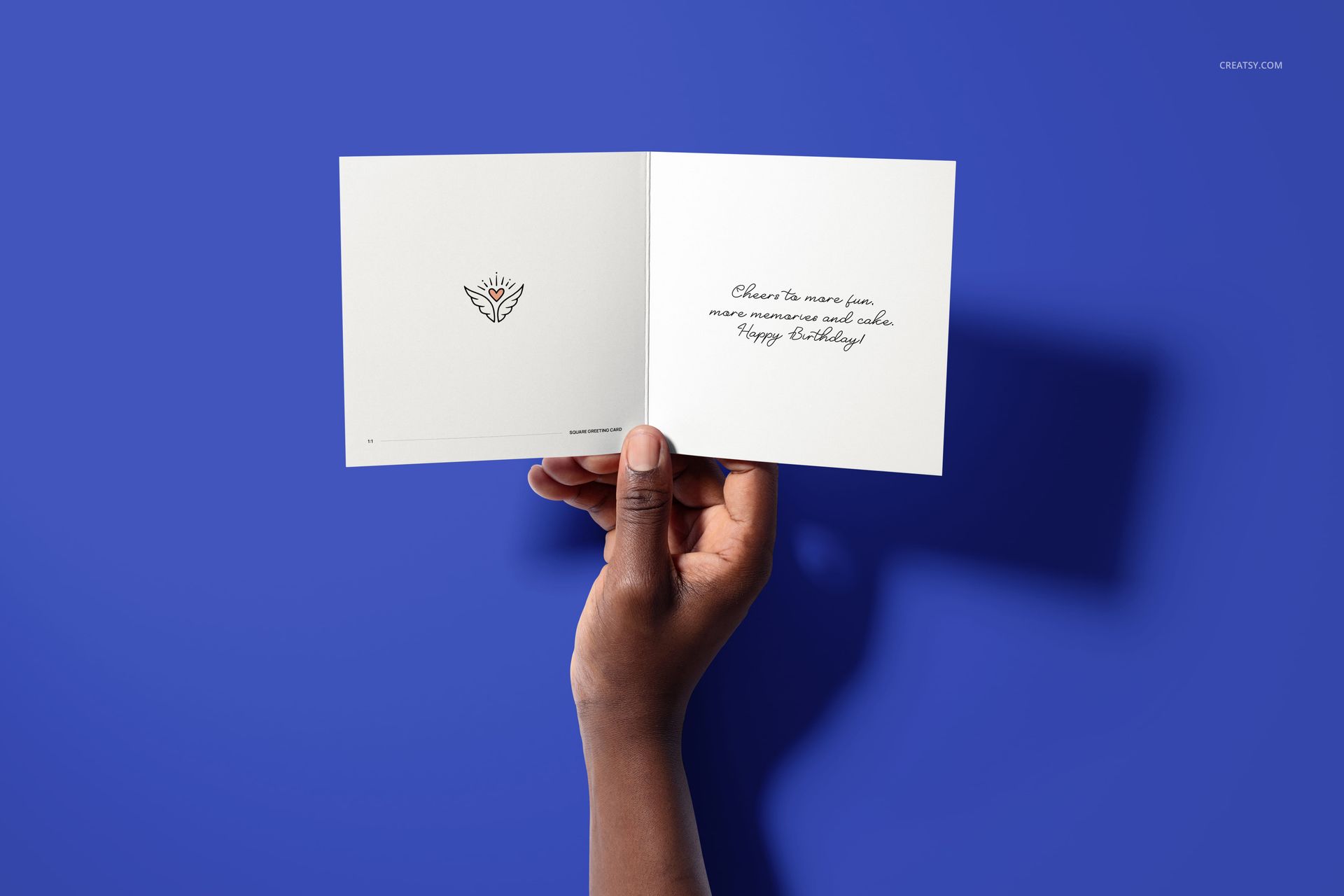 Open square folded greeting card mockup featuring handwritten message and simple graphic, held by hand on blue surface.