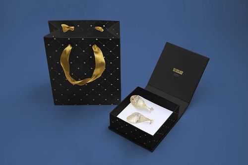 Jewelry box mockup set featuring an open black box with gold foil stamp and matching patterned gift bag on a blue surface.