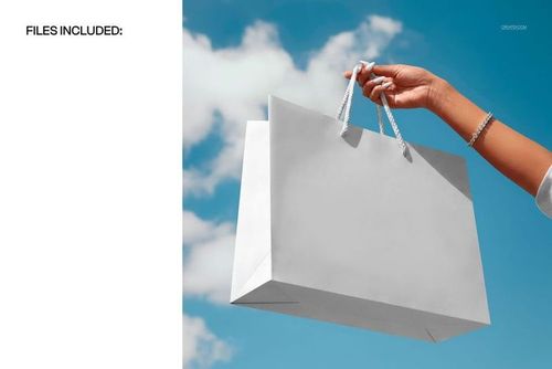 PSD mockup showing a white paper shopping bag held by hand outdoors, with blue sky and scattered clouds behind.