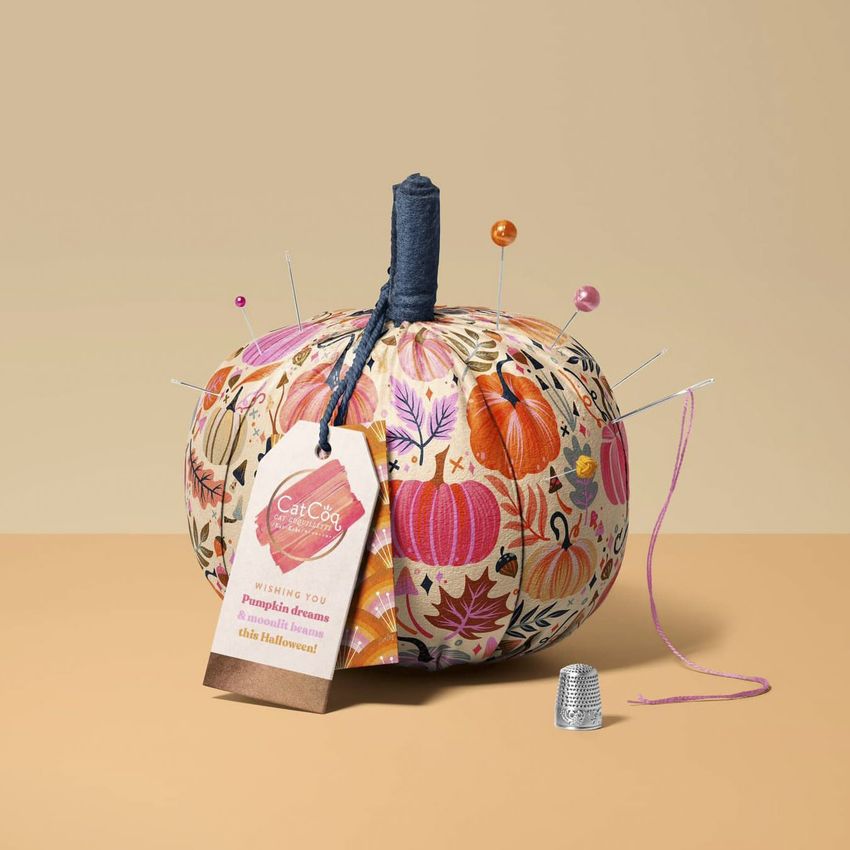 Fabric pumpkin mockup featuring a round, patterned pumpkin with sewing pins, a needle, a pink thread, and a thimble on a beige surface.