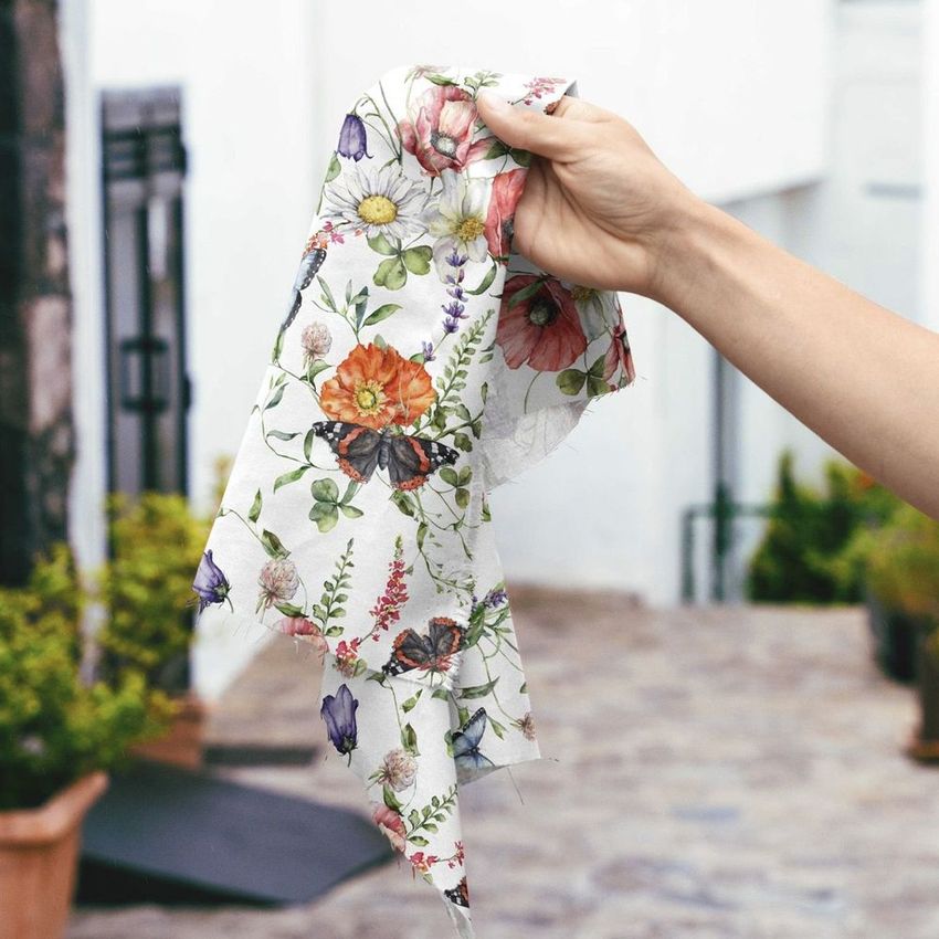 Hand holding a floral fabric sample against a rustic urban backdrop with stone pavement and potted plants in the background.