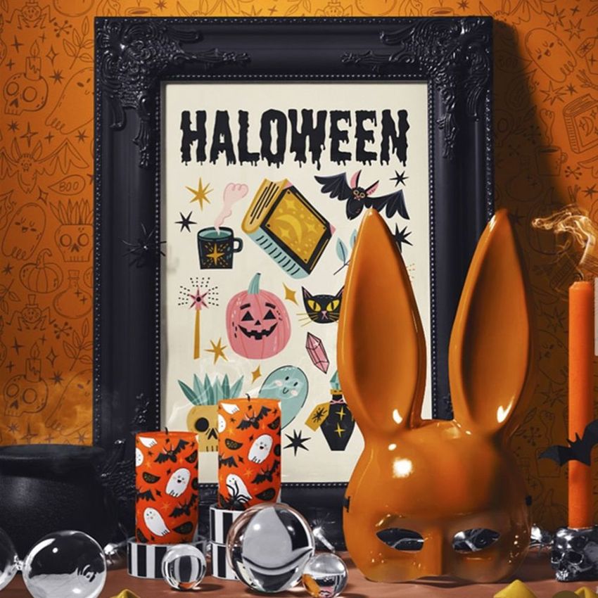 Halloween art print mockup in an ornate black frame, featuring festive illustrations and surrounded by themed props on an orange background.