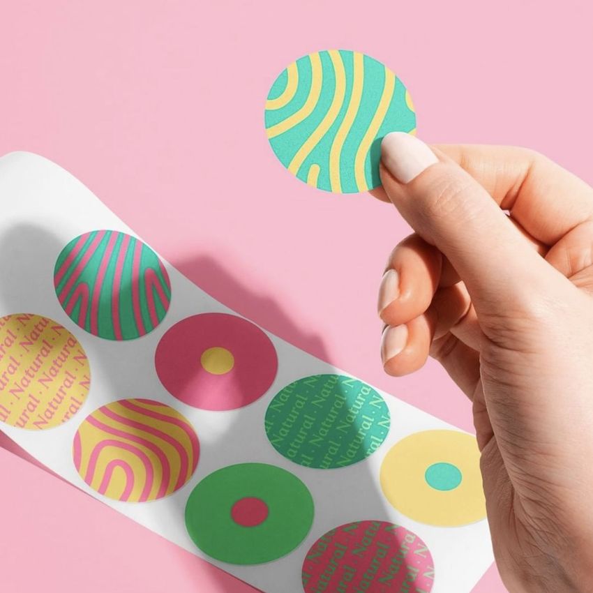 Hand holding a round, colorful sticker above a sheet of assorted circular stickers with abstract patterns on a pink background.