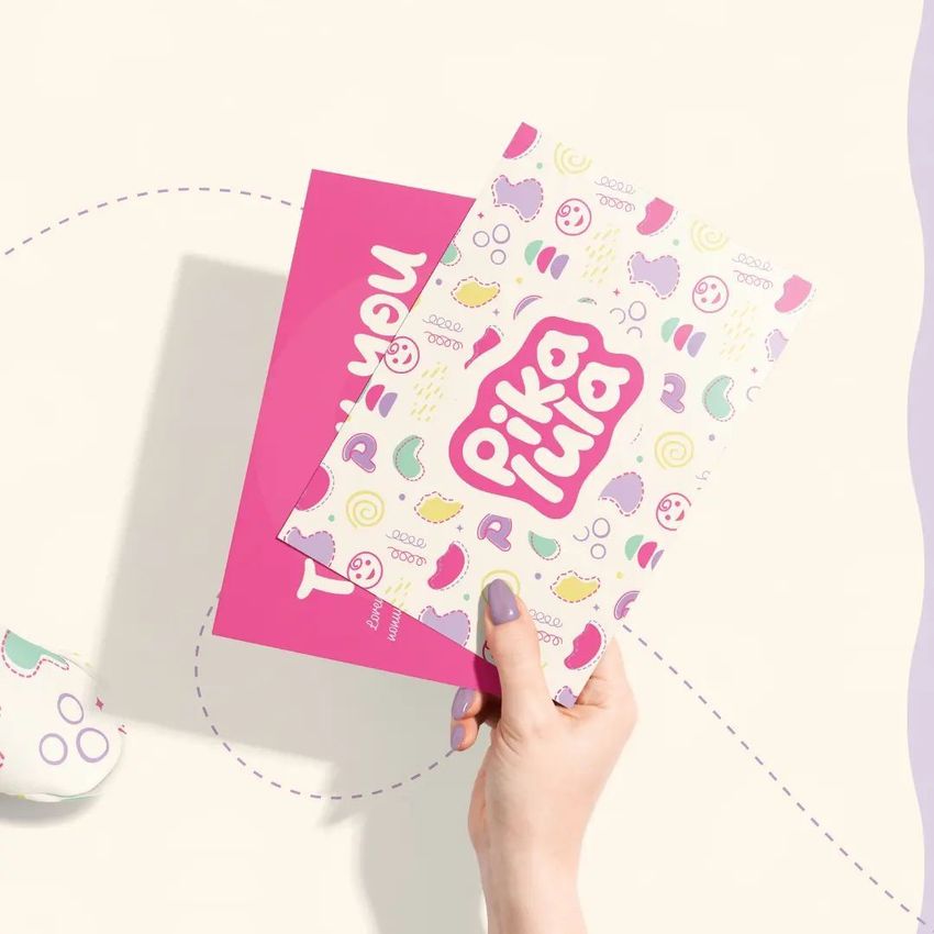 Hand holding two stationery cards, one with a colorful abstract pattern and bold text, the other solid pink with white text, on a light background.