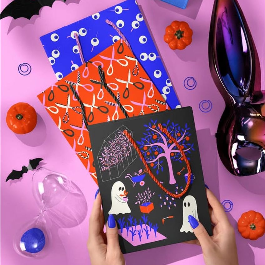 Hands holding a Halloween paper gift bag with ghost and tree illustrations, surrounded by pumpkins, bats, and decorative props on a pink surface.