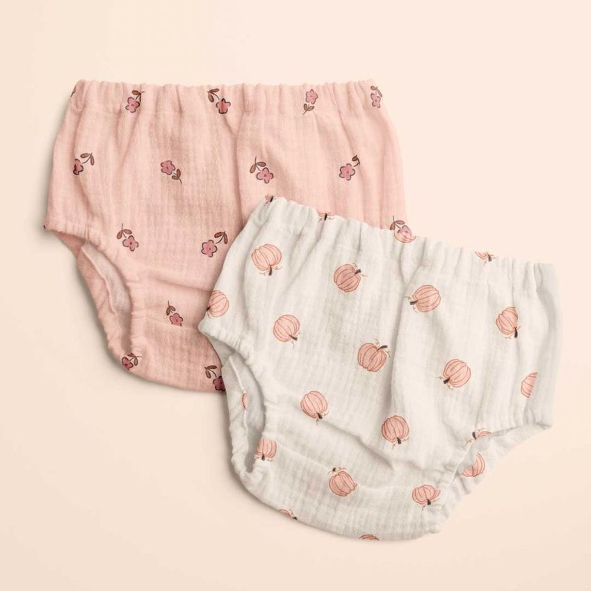 Flat lay of baby muslin pants mockup featuring a pink pair with floral designs and a white pair with peach motifs on a neutral backdrop.