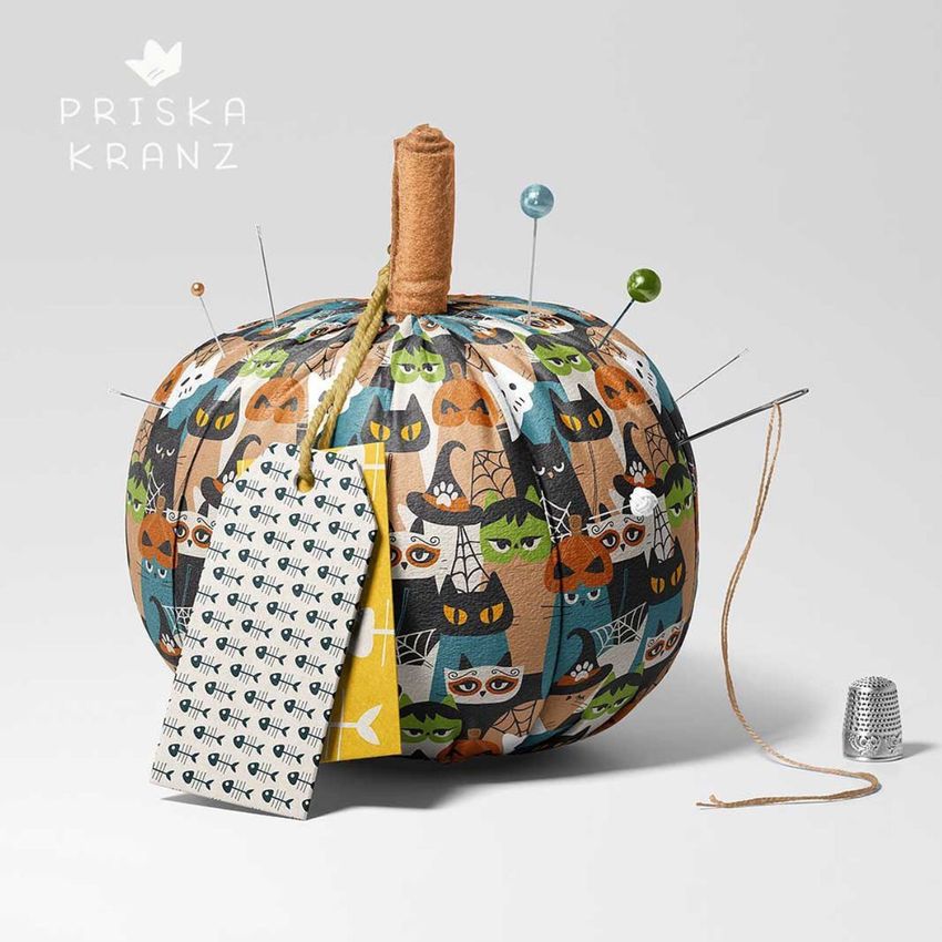 Fabric pumpkin mockup decorated with a colorful Halloween cat pattern, used as a pincushion with pins and a thimble nearby.