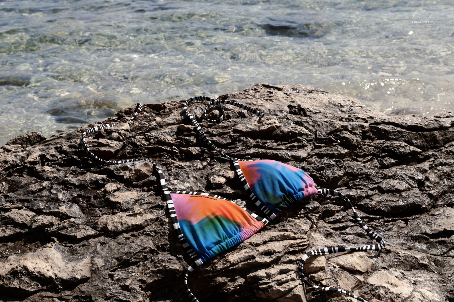 Vibrant bikini mockup featuring multicolored fabric and black-and-white ties, laid out on seaside rocks outdoors.