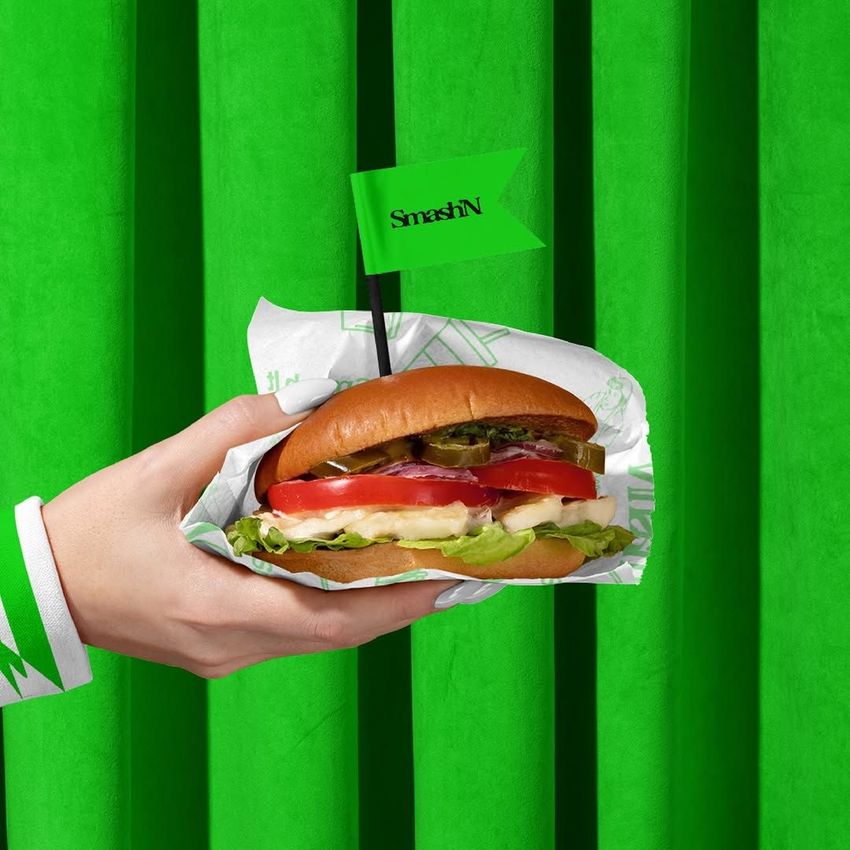 Hand holding a vegan burger wrapped in branded paper with a small green flag, set against a bright green background.