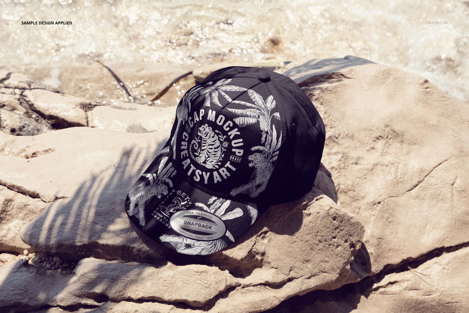 Front view of a classic snapback cap mockup featuring bold white graphics, placed on rocky terrain by the water.