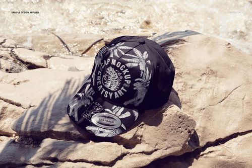 Front view of a classic snapback cap mockup featuring bold white graphics, placed on rocky terrain by the water.